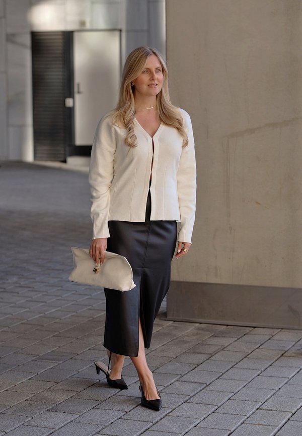 Femme aux longs cheveux blonds portant une veste blanche, une jupe midi en cuir noire, des talons noirs et tenant une pochette beige, marchant dans une rue pavée.