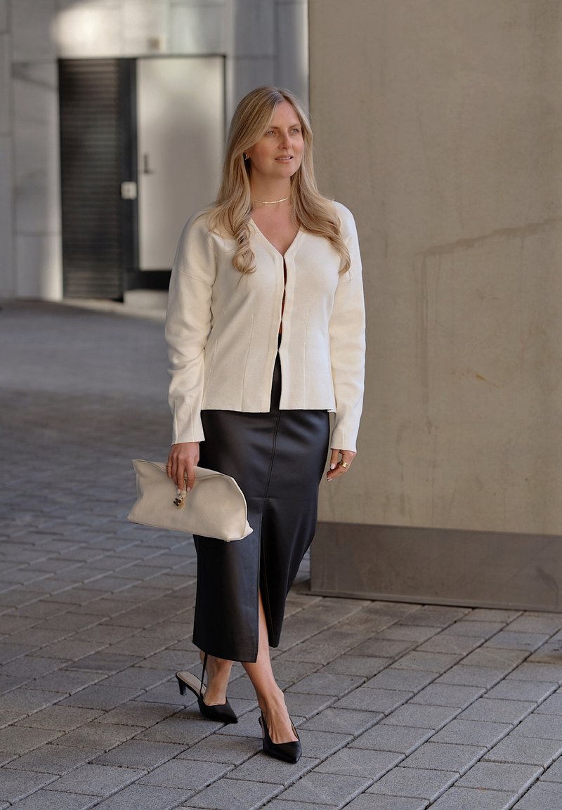 Femme aux longs cheveux blonds portant une veste blanche, une jupe midi en cuir noir, des talons noirs et tenant une pochette beige, marchant dans une rue pavée.