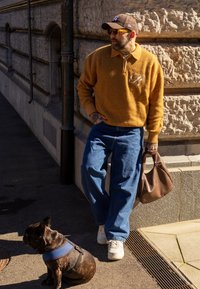 Pull tricoté orange avec fermeture éclair, jean bleu, baskets blanches, sac à main marron. Chien noir avec harnais bleu assis à proximité, mur en pierre en arrière-plan.
