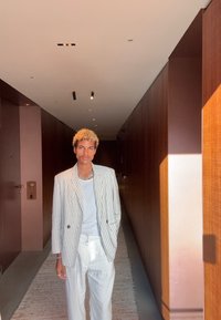 Νεαρός με σγουρά ξανθά μαλλιά, που φοράει ριγέ σακάκι, άσπρο πουκάμισο και άσπρο παντελόνι, στέκεται σε έναν ηλιόλουστο σύγχρονο διάδρομο.