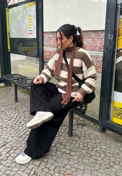 Mujer con un suéter a rayas marrón y blanco, pantalones negros y zapatillas blancas sentada en una parada de autobús con un mapa de Berlín detrás de ella.