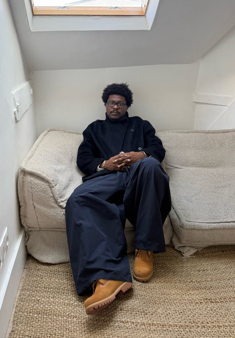 Άνδρας με γυαλιά που φοράει μαύρο παλτό και φαρδιά μπλε παντελόνια κάθεται σε μια μπεζ καναπέ κάτω από ένα μικρό παράθυρο φωταγωγού.