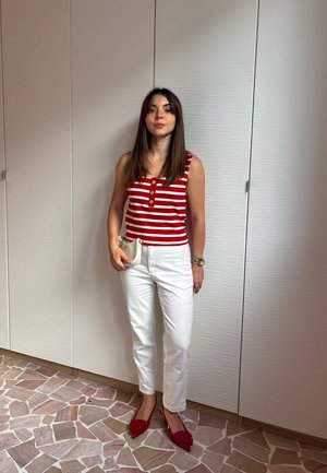 Top sin mangas a rayas rojas y blancas con botones, pantalones blancos y zapatos planos rojos de punta. Pequeño bolso blanco, de pie sobre azulejos de suelo estampados.