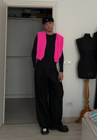 Pantalones oversized negros, camisa negra de manga larga ajustada, suéter rosa brillante sobre los hombros, zapatos de goma negros con un acento plateado.