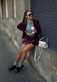 Blazer bordeaux, maglione grigio a maglia, mini gonna in pelle bordeaux, calze grigie, mocassini neri lucidi, piccola borsa a tracolla bianca, in piedi contro un muro di pietra.