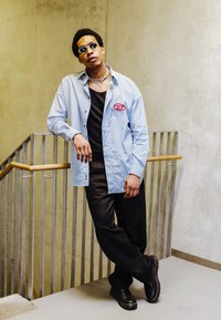 Camicia a righe blu e bianche con un'applicazione rossa, canotta nera, pantaloni larghi neri, occhiali da sole tondi e catena d'argento.