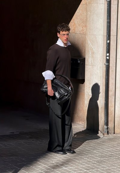 Joven con gafas, suéter negro sobre camisa blanca, sosteniendo una gran bolsa negra, de pie sobre un pavimento de baldosas cerca de un muro de piedra bajo la luz del sol.