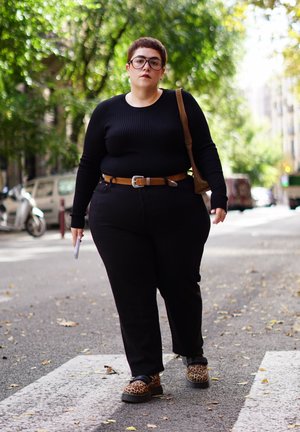 Persona con cabello corto y gafas camina por un paso de peatones en la ciudad, vistiendo ropa negra, cinturón marrón, zapatos estampados de leopardo y llevando un bolso marrón.