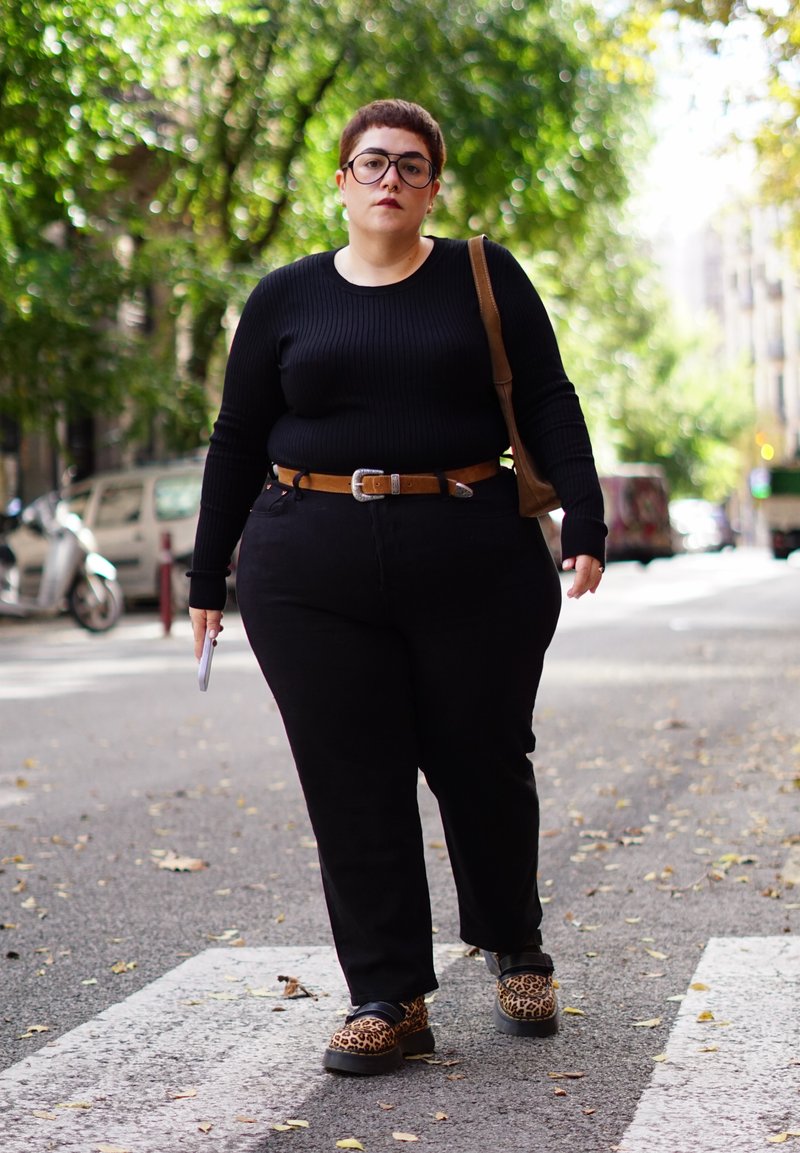 Person mit kurzem Haar und Brille geht an einem Zebrastreifen in der Stadt. Sie trägt ein schwarzes Outfit, einen braunen Gürtel, Leopardenmuster-Schuhe und trägt eine braune Tasche.