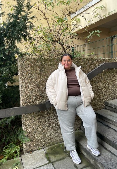 Chaqueta de crema acolchada sobre una blusa marrón, pantalones de chándal grises y zapatillas blancas. De pie al lado de una pared de hormigón texturizada con follaje.