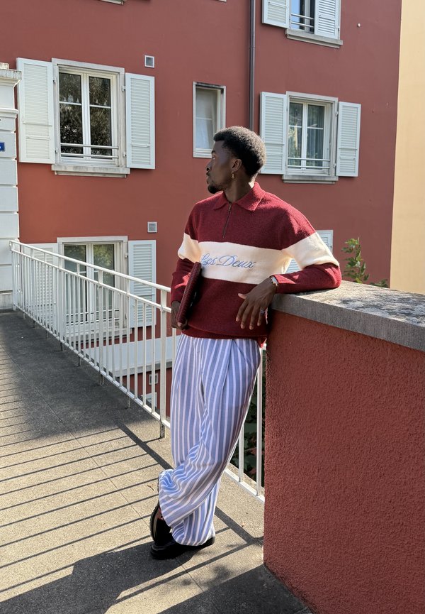 Homme portant un pull rouge et blanc et un pantalon rayé bleu s'appuie sur un mur rouge sur un balcon avec des volets blancs derrière lui.