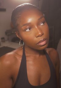 Portret osoby o gładkiej, promiennej cerze, ubranej w czarny top na szyję, z dużymi kołowymi kolczykami i ciemnymi, zaczesanymi do tyłu włosami.