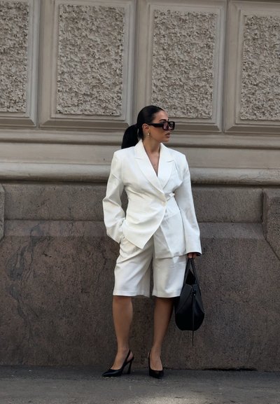 Mujer de pie contra una pared de piedra texturizada, vestida con blazer blanco de gran tamaño y shorts, tacones altos negros, gafas de sol y sosteniendo un bolso negro.