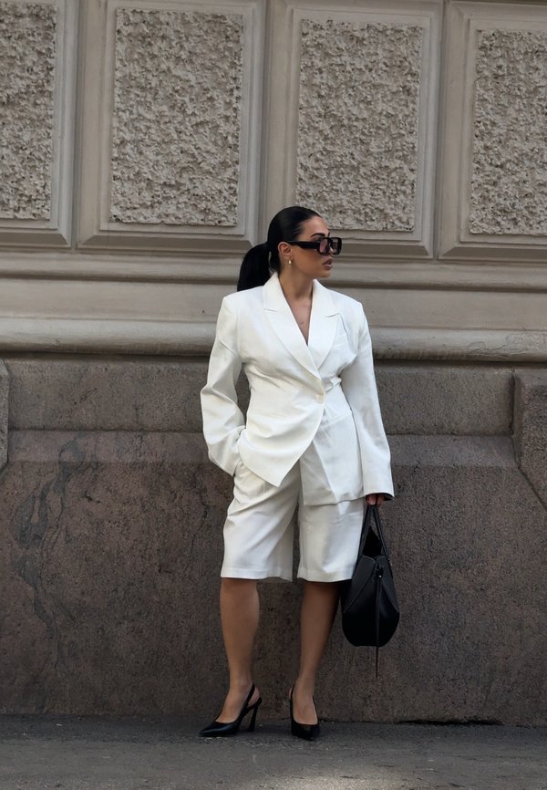 Kvinde står op ad en struktureret stenvæg iført hvid oversized blazer og shorts, sorte højhælede sko, solbriller og holder en sort håndtaske.