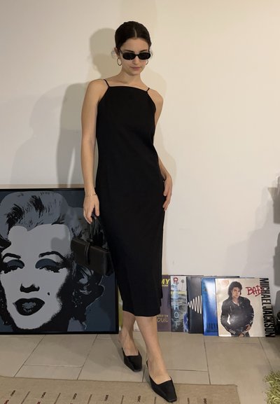Mujer con vestido negro y gafas de sol sosteniendo un bolso negro, de pie frente a una pared con arte enmarcado de Marilyn Monroe y discos de vinilo.