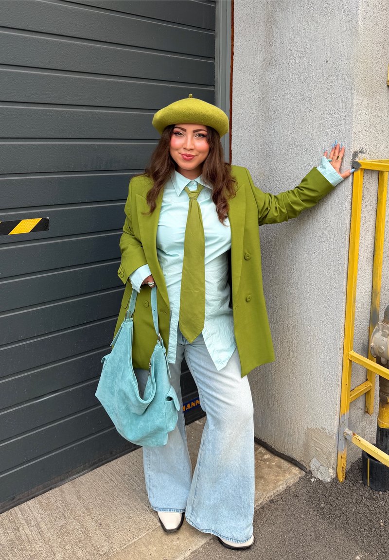 Woman wearing a green beret, green blazer, light green shirt, and tie, holding a blue bag while standing by a gray door and concrete wall.