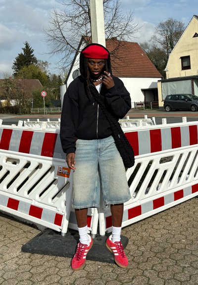 Una persona con una sudadera negra y pantalones cortos de mezclilla lleva una boina roja, calcetines blancos y zapatillas rosas, de pie junto a barreras de construcción rojas y blancas.