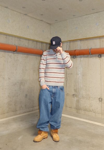 Hombre con suéter gris a rayas, jeans azules holgados, botas color canela y gorra negra, de pie en un sótano de concreto con tuberías naranjas, ajustando el ala de la gorra con la mano.