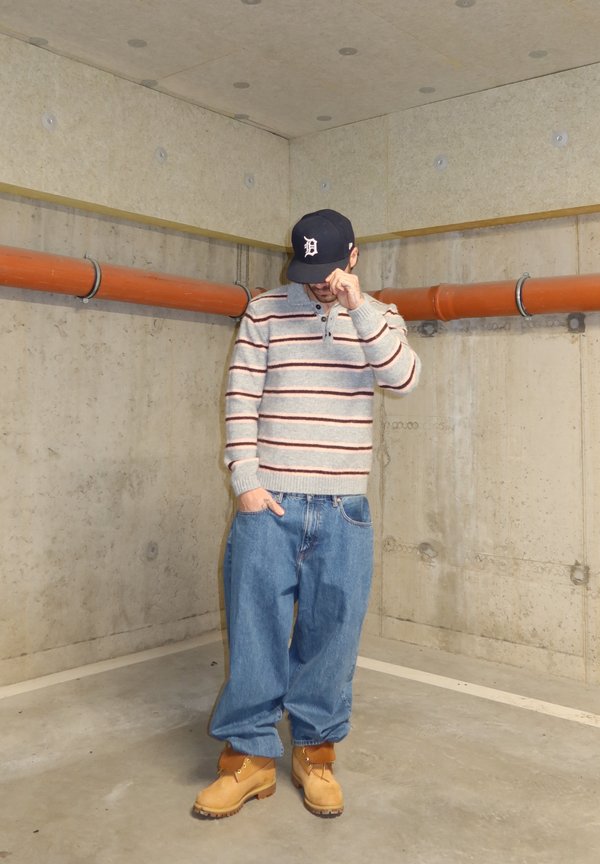 Mies, joka pukeutunut harmaaraidalliseen neulepuseroon, väljihin sinisiin farkkuihin, vaaleanruskeisiin saappaisiin ja mustaan lippalakkiin, seisoo betonisessa kellarissa, jossa on oranssit putket, ja säätää lippalakin lippaa kädellään.