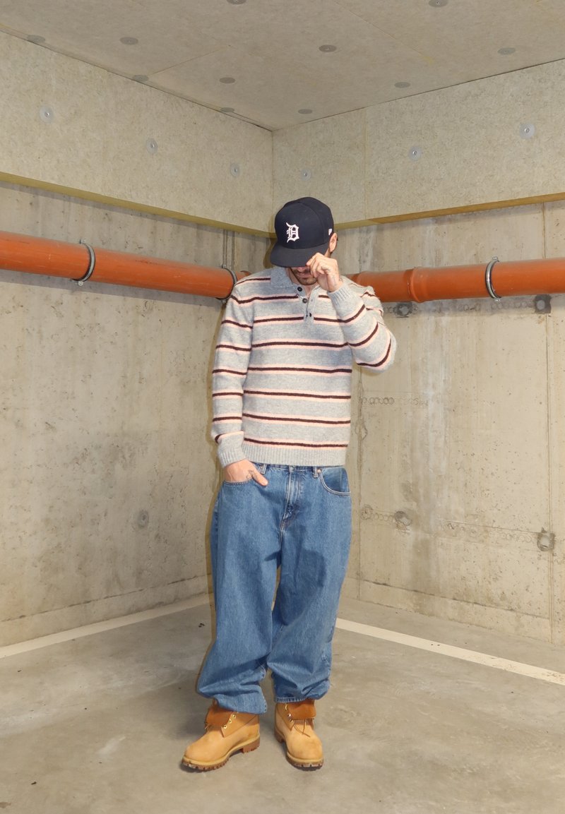 Uomo con maglione a righe grigio, jeans blu larghi, stivali marroni chiari e cappellino nero che si trova in un seminterrato in cemento con tubi arancioni, mentre si sistema la visiera del cappellino con la mano.