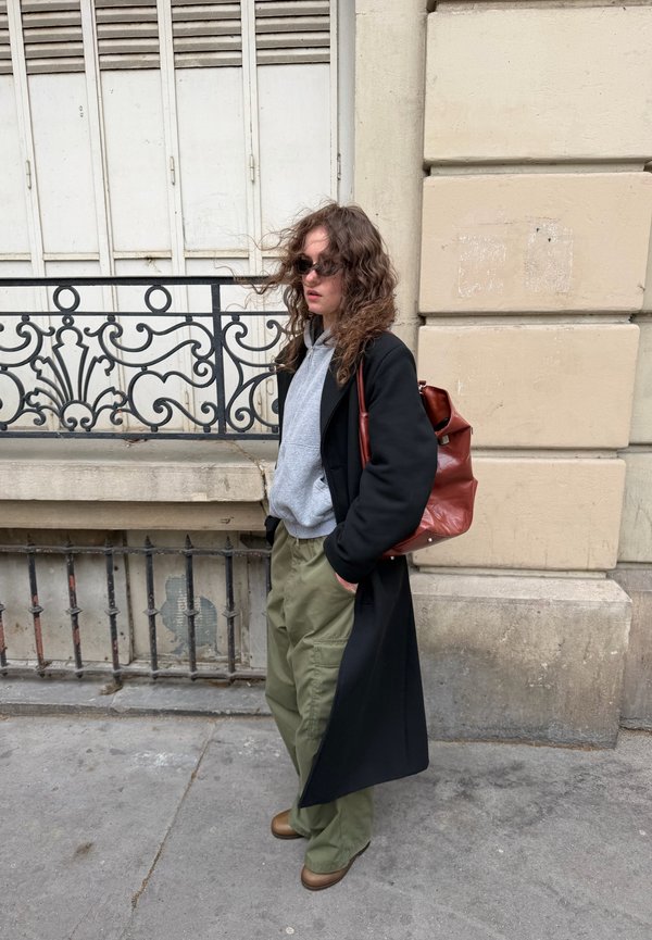 Frau mit lockigem Haar, die eine Sonnenbrille, einen grauen Hoodie, einen schwarzen Mantel, grüne Cargohosen, braune Stiefel trägt und einen braunen Lederrucksack auf einem Bürgersteig in der Stadt trägt.