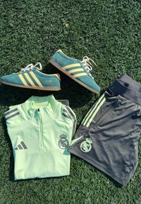 Adidas sportska odjeća koja se sastoji od majice s patentnim zatvaračem u mint zelenoj boji, crnih šortseva s zelenim detaljima i zeleno-žutih tenisica s tri pruge.