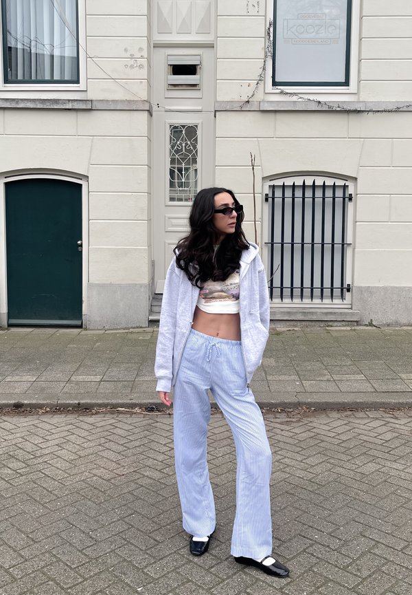 Balts džemperis ar kapuce virs rakstainā piegriezuma topa, apvienots ar gaiši ziliem svītrainiem plaša griezuma biksēm un melnām kurpēm, stāvot uz bruģētas ielas.