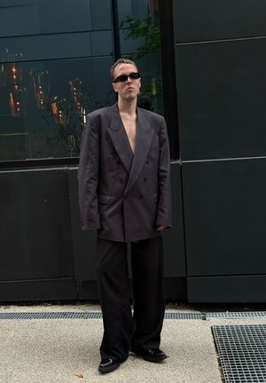 Übergroße, doppelt geknöpfte dunkelgraue Blazer mit breiten Revers, kombiniert mit lockeren schwarzen Hosen und schwarzen Schuhen. Sonnenbrillen runden den Look ab.