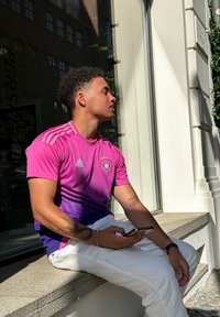 Ρόδινο και μοβ gradient Adidas μπλούζα με τρία αστέρια και λογότυπο, σε συνδυασμό με λευκά παντελόνια. Άτομο που κρατά ένα τηλέφωνο, καθισμένο σε ένα παράθυρο.