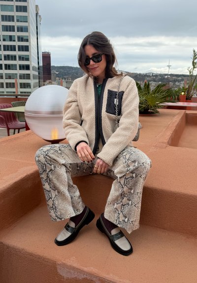 Mujer con gafas de sol, chaqueta de forro polar y pantalones con estampado de serpiente, sentada en unas escaleras de la azotea con la ciudad y plantas detrás de ella.