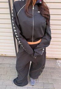 Personne portant un sweat à capuche noir zippé et un pantalon ample avec des bandes blanches à logo sur les manches, debout sur un trottoir pavé.