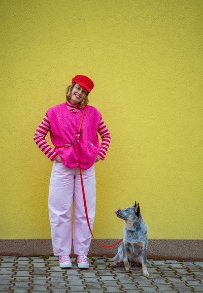 Mujer con ropa rosa y gorra roja sonriendo, sosteniendo una correa roja atada a un perro blue heeler sentado contra una pared amarilla.