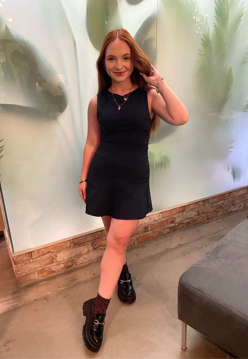 Schwarzes ärmelloses Kleid mit tailliertem Oberteil und ausgestellt Rock. Tragen mit schwarzen klobigen Schuhen und bordeauxroten gemusterten Socken. Silberne Schmuckakzente.