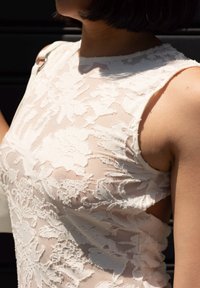 Biała koronkowa bluzka z kwiatowymi wzorami, bez rękawów i przezroczystą teksturą. Dekolt jest okrągły z otwartymi bokami.