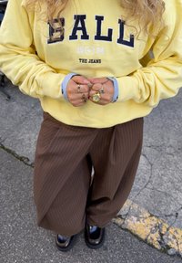 Persona che indossa una felpa gialla con la scritta "BALL", pantaloni larghi marroni a righe sottili, scarpe nere e diversi anelli, incluso un anello con orologio d'oro sulla mano sinistra.