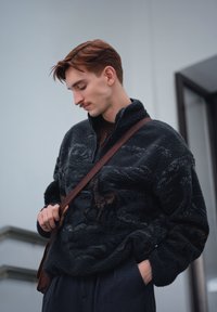 Pull polaire en peluche noire avec un col montant et une fermeture éclair. Présente une surface texturée et une bretelle marron sur l'épaule. Pantalon foncé.