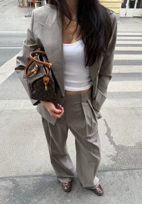 Costum gri croit cu pantaloni evazați și o geacă structurată. Top alb subțire; ținând o geantă de mână cu un model maro și bej.