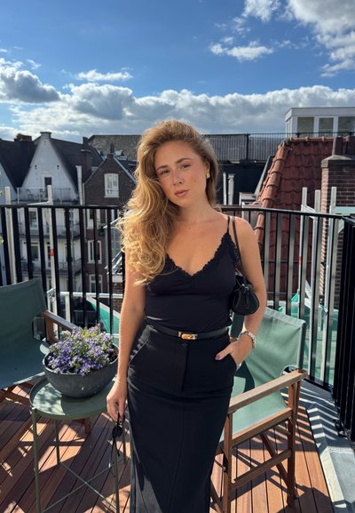 Top negro sin mangas con detalles de encaje, falda negra de tiro alto, pendientes dorados y pequeño bolso negro. Terraza de madera con plantas verdes.