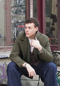 Casaco de lã verde-azeitona com grandes lapelas, vestido sobre um colete às riscas. Calças de ganga azuis e pulseira prateada. O fundo apresenta fotografias a preto e branco.