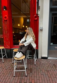 Haut blanc à manches longues, pantalon ample marron et baskets blanches. Tabourets en bois avec pieds métalliques autour d'une devanture rouge. Fond en pavés de briques.