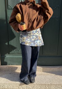 Person iført en brun jakke, blomster nederdel over mørke jeans, holder en gul clutch og en takeaway kaffekop, står ved en grøn dør.