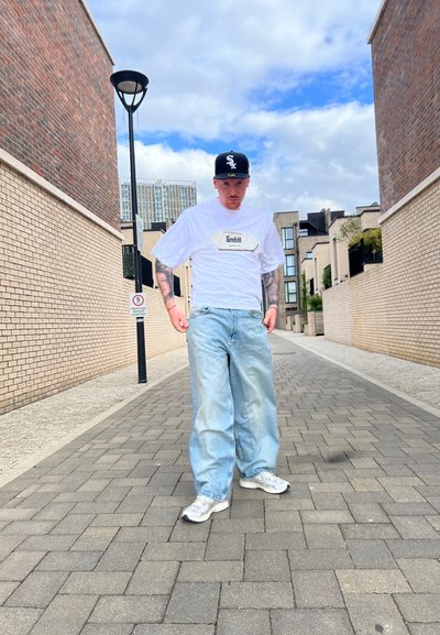 Camiseta blanca oversized con logo "GmbH", pantalones vaqueros de pierna ancha en azul claro y zapatillas grises, colocados en un callejón pavimentado entre edificios de ladrillo.