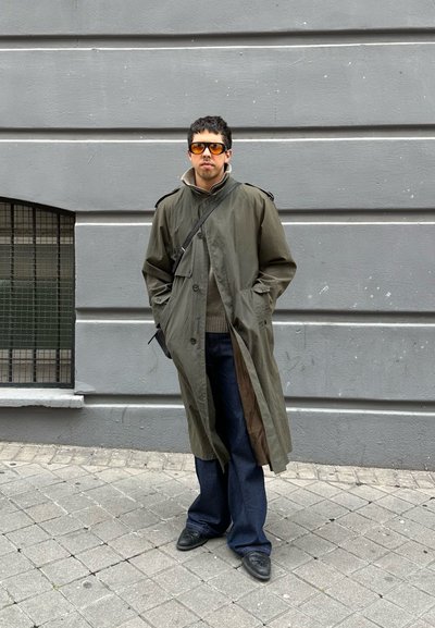 Hombre con abrigo tipo gabardina verde oliva y gafas de sol anaranjadas de pie en una acera con baldosas frente a una pared gris.