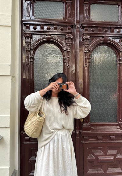 Suéter de punto crema y falda blanca fluida, sosteniendo una cámara, con un bolso de mimbre beige, posando frente a puertas marrones ornamentadas.