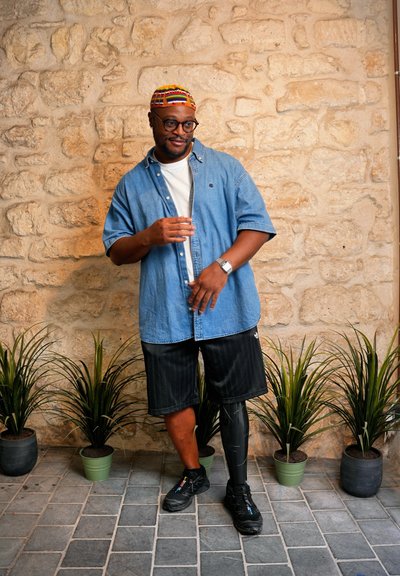 Camisa de mezclilla azul de manga corta sobre una camiseta blanca, pantalones cortos negros a rayas, zapatillas negras y una pierna prostética negra con detalles en blanco.