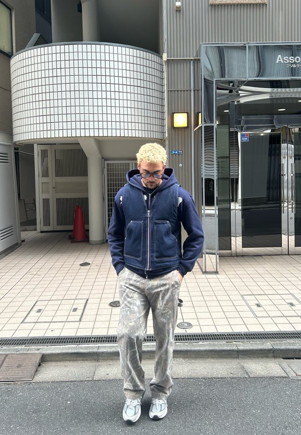 Jeansweste über einem marineblauen Hoodie, kombiniert mit hellgrauen Camouflage-Hosen und grauen Sneakers. Steht auf einem gefliesten Gehweg in der Nähe eines Glasgebäudes.