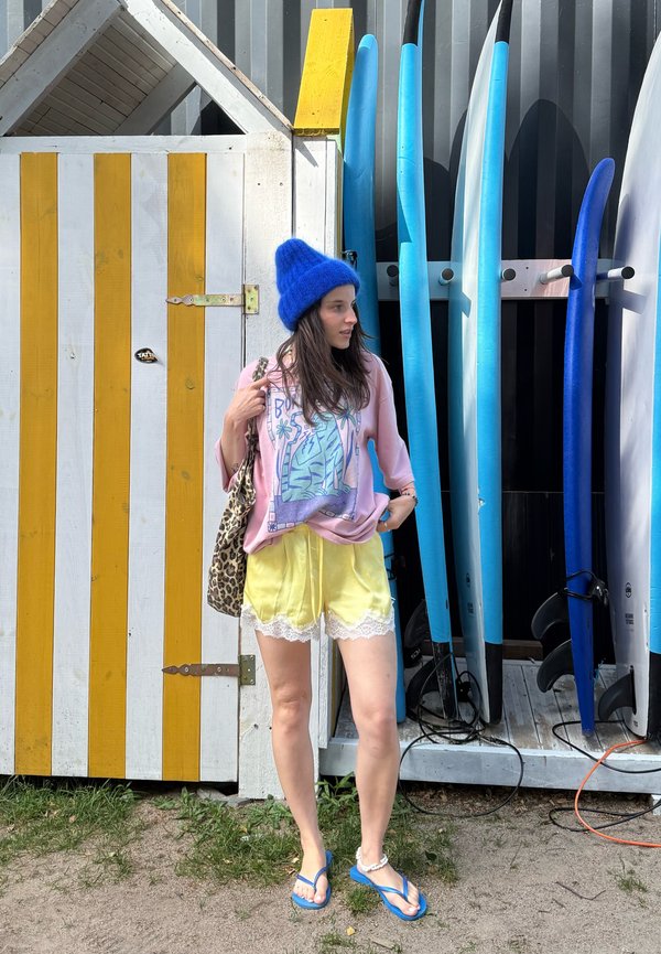 Młoda kobieta w jasnych, codziennych ubraniach i niebieskiej czapce stoi obok pasiastego domku na plaży i rzędu stojących pionowo desek surfingowych.