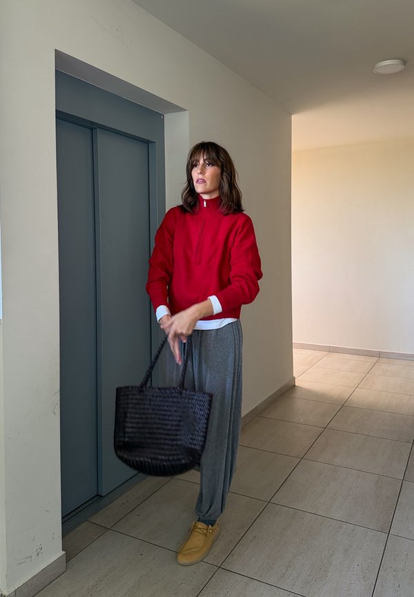 Une femme vêtue d’un haut rouge, d’un pantalon gris et de chaussures beiges tient un sac noir tissé à côté d’un ascenseur.