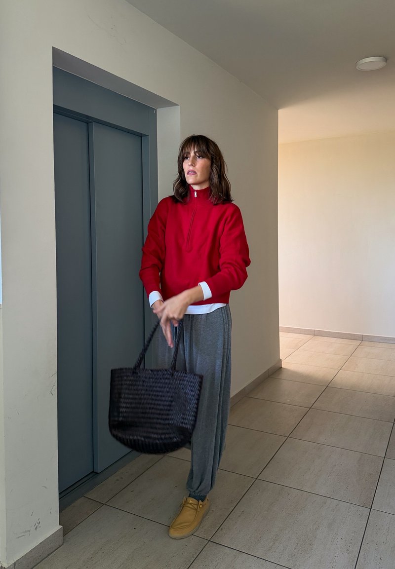 Une femme vêtue d’un haut rouge, d’un pantalon gris et de chaussures beiges tient un sac noir tissé à côté d’un ascenseur.