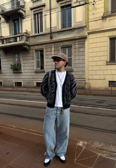 Cárdigan de patrón negro y gris sobre una camiseta blanca, combinado con jeans sueltos de color azul claro y zapatos blanco y negro. Los accesorios incluyen una cadena y una gorra.